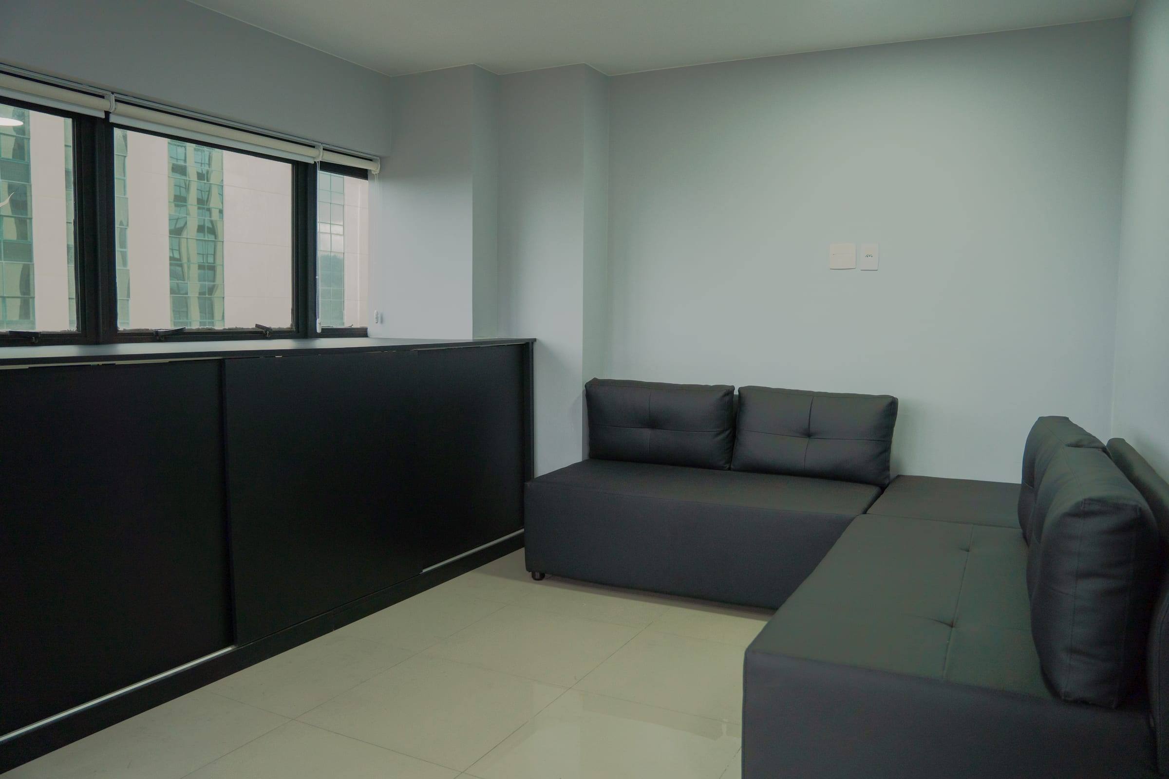 Sala de estar com sofá estofado em cinza-escuro e janelas para os prédios da Barra da Tijuca