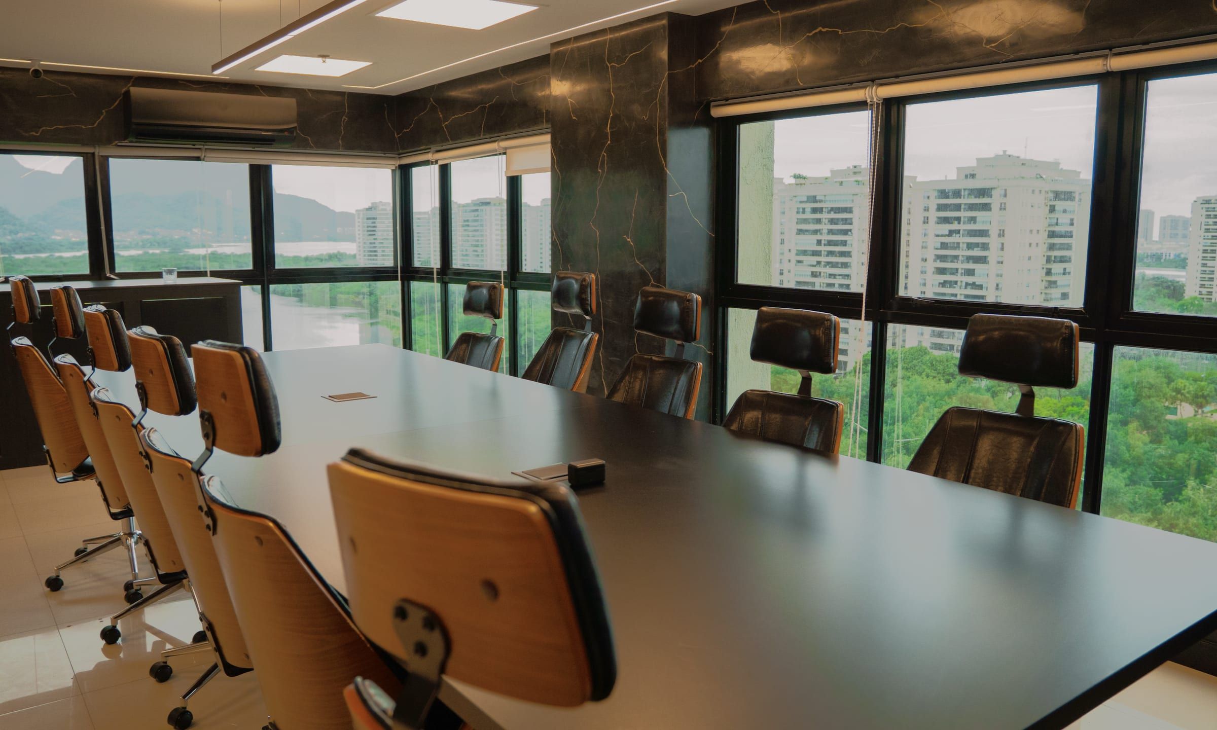 Sala de reunião com mesa longa, cadeiras em couro e madeira e janelas para as montanhas e os prédios da Barra da Tijuca
