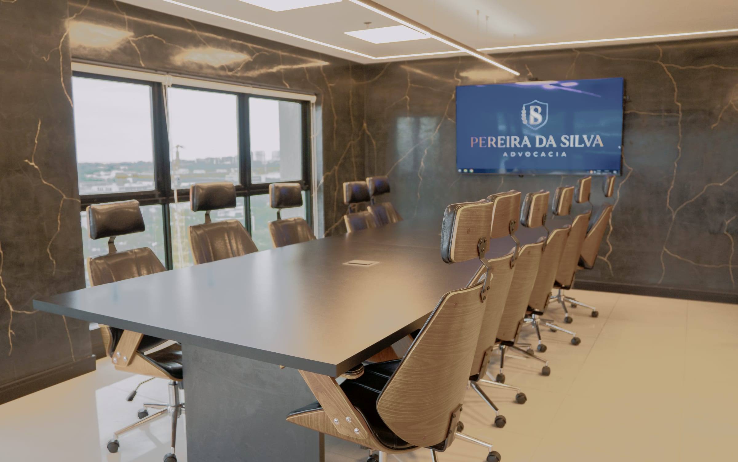 Sala de reunião com mesa longa, cadeiras de couro e madeira, janelas para a Barra da Tijuca e tela com o logotipo do escritório