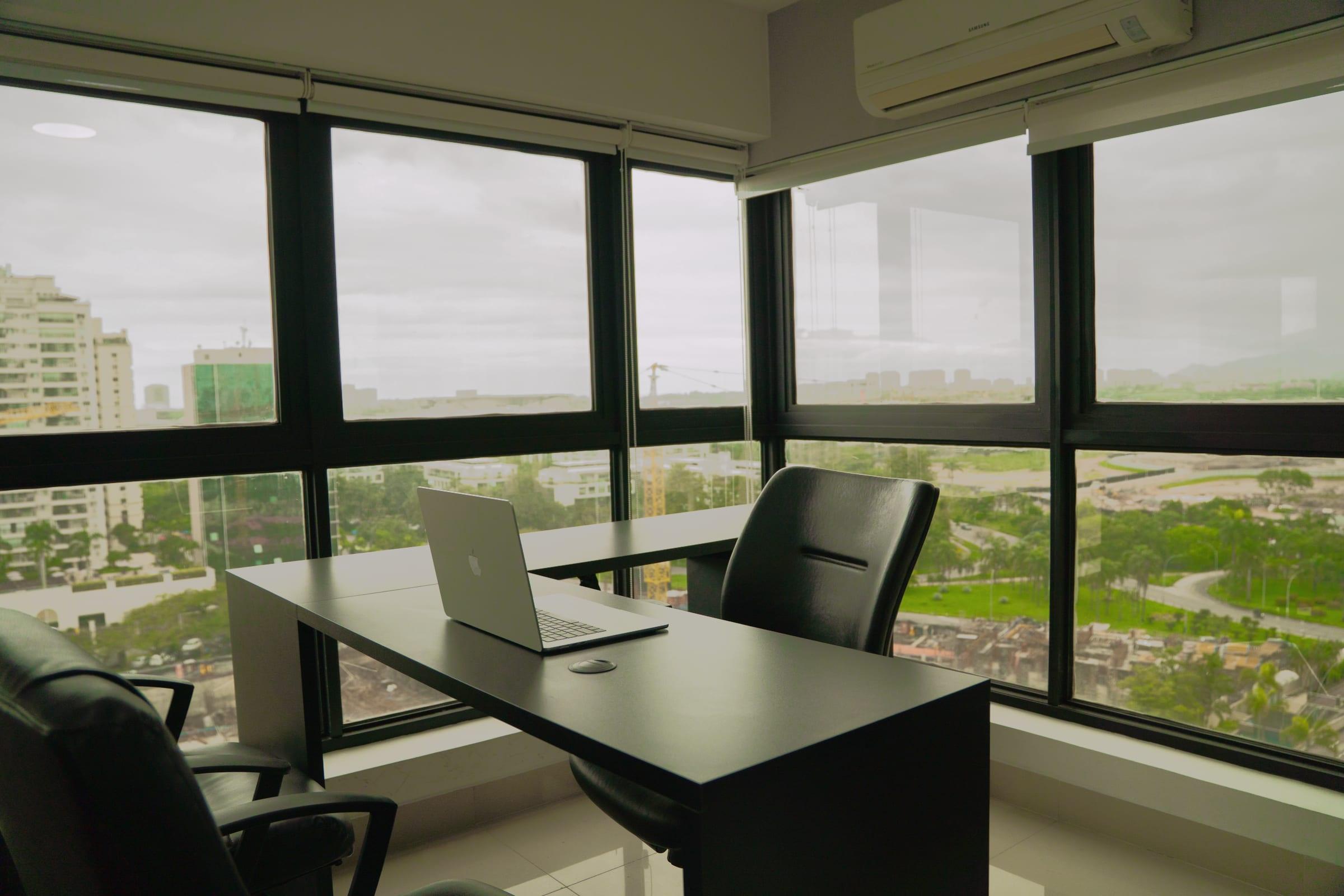 Sala de atendimento com mesa escura, cadeira executiva e janelas de canto voltadas para a vegetação e os prédios da Barra da Tijuca