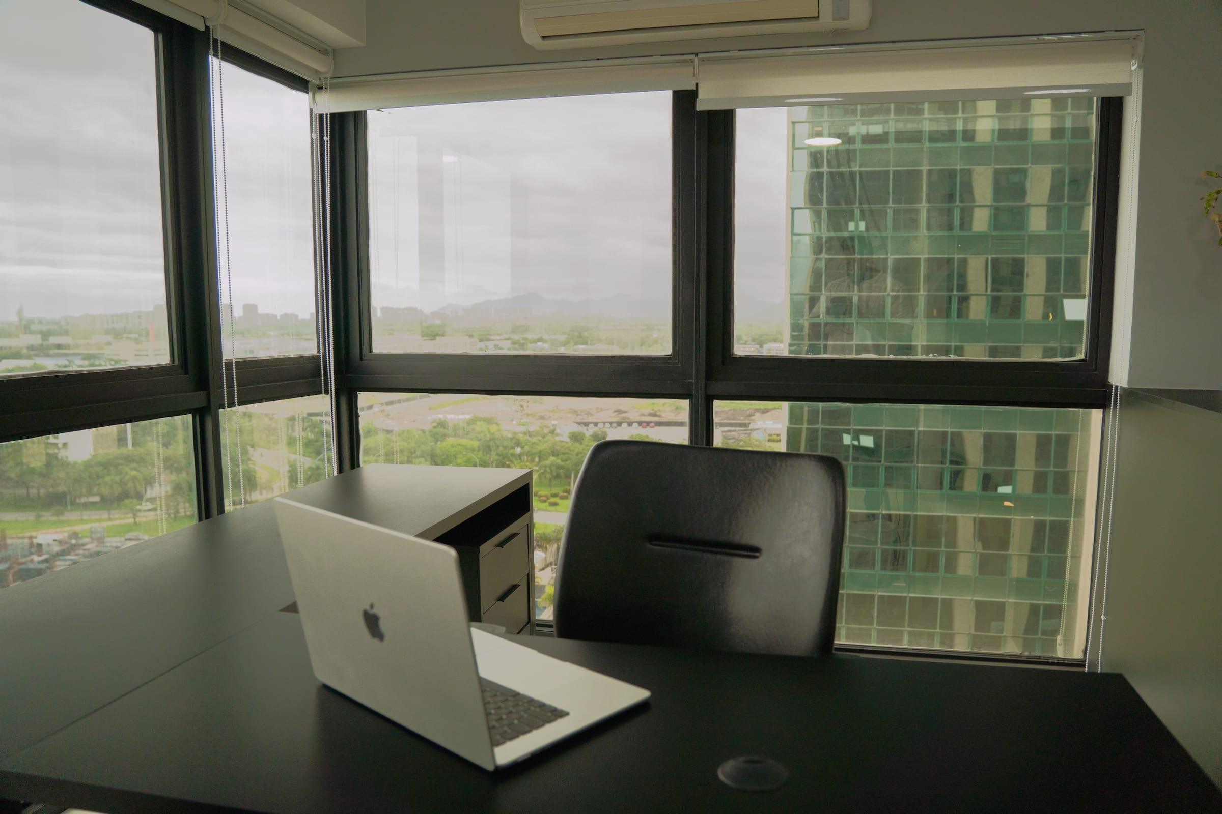 Sala de atendimento com mesa escura, notebook e janelas amplas voltadas para os prédios e a vegetação da Barra da Tijuca