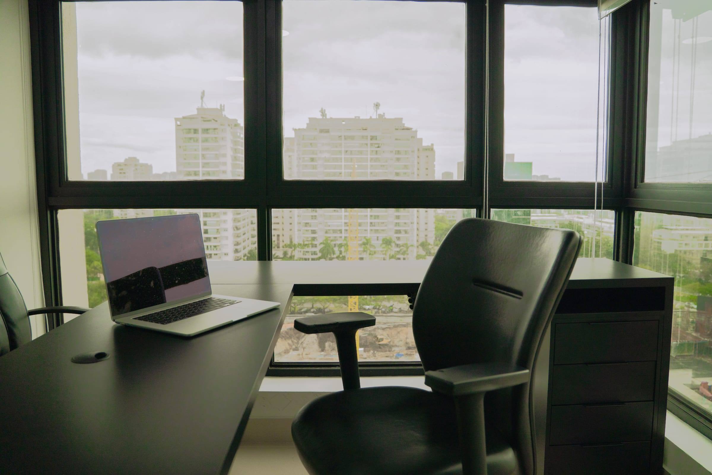 Sala de atendimento com mesa escura, notebook e janelas amplas voltadas para os prédios da Barra da Tijuca