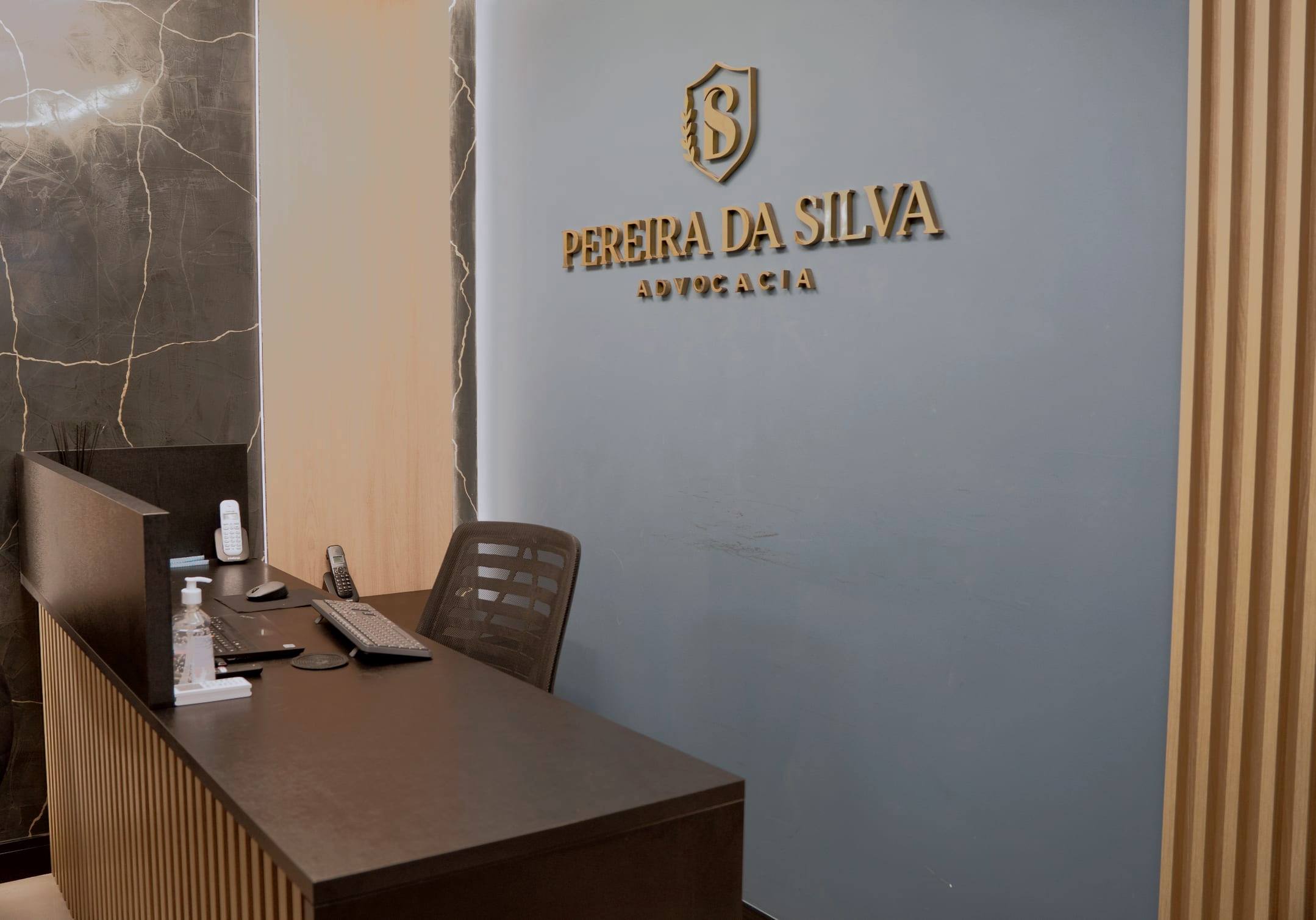 Balcão da recepção em madeira escura diante da parede azul-clara com o letreiro Pereira da Silva Advocacia em relevo dourado